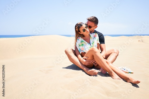 Young beautiful couple smiling happy and confident. Sitting with smile on face hugging and kissing at the beach