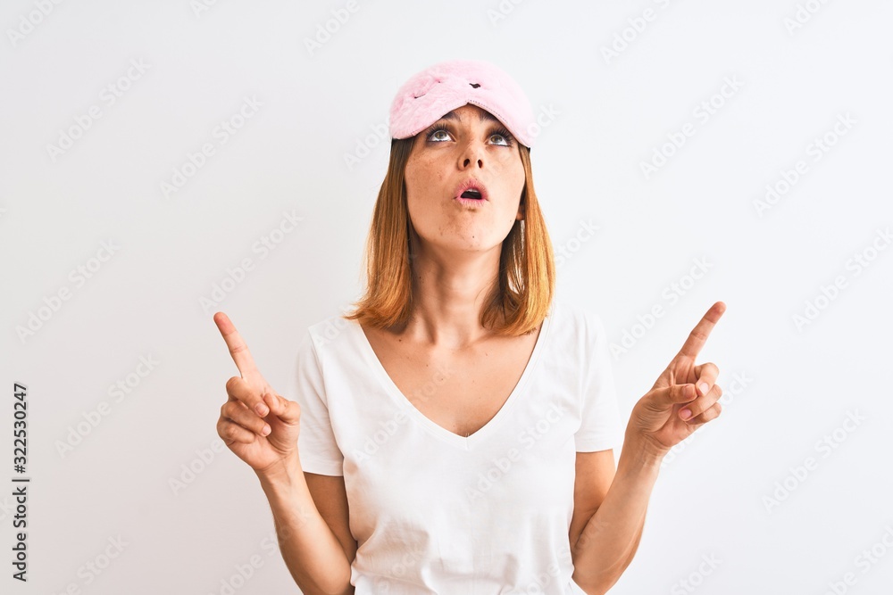 Beautiful redhead woman wearing sleeping mask over isolated background amazed and surprised looking up and pointing with fingers and raised arms. Beautiful redhead woman wearing sleeping mask over isolated background amazed and surprised looking up and pointing with fingers and raised arms.