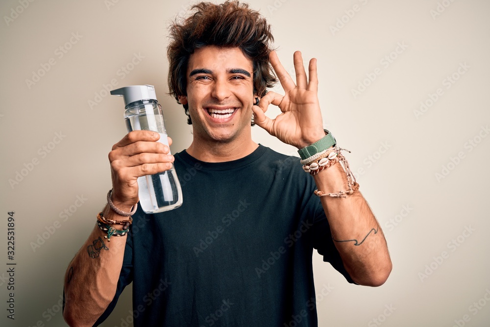 Young handsome man holding bottle of water standing over isolated white background doing ok sign with fingers, excellent symbol Young handsome man holding bottle of water standing over isolated white background doing ok sign with fingers, excellent symbol