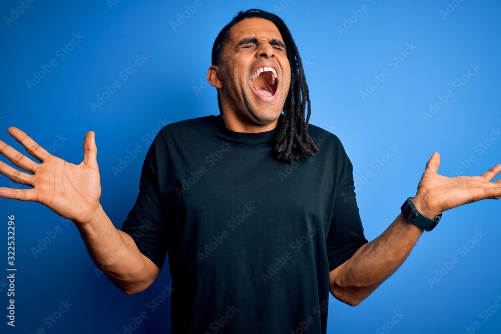 Fototapeta premium Young handsome african american afro man with dreadlocks wearing casual t-shirt celebrating mad and crazy for success with arms raised and closed eyes screaming excited. Winner concept
