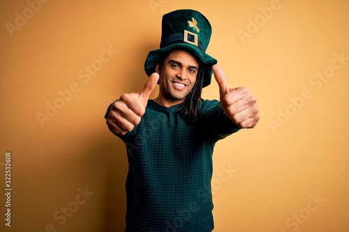 Young african american man wearing green hat with clover celebrating saint patricks day approving doing positive gesture with hand, thumbs up smiling and happy for success. Winner gesture.