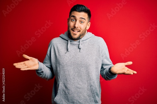 Young handsome sportsman wearing sweatshirt standing over isolated red background clueless and confused expression with arms and hands raised. Doubt concept.