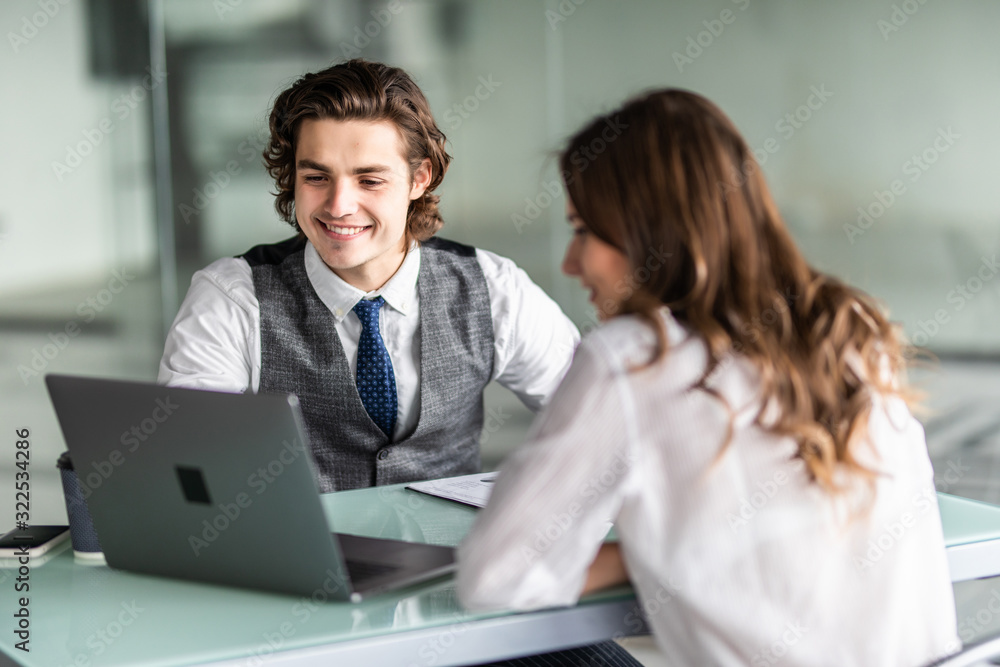 Beautiful young business woman and handsome businessman in formal suits are using a laptop, discussing documents and smiling while working in office Beautiful young business woman and handsome businessman in formal suits are using a laptop, discussing documents and smiling while working in office