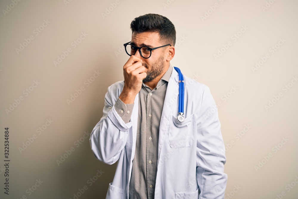 Young doctor man wearing glasses, medical white robe and stethoscope over isolated background smelling something stinky and disgusting, intolerable smell, holding breath with fingers on nose. Young doctor man wearing glasses, medical white robe and stethoscope over isolated background smelling something stinky and disgusting, intolerable smell, holding breath with fingers on nose.