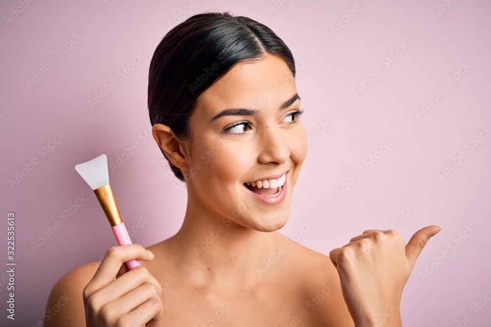 Young beautiful girl applying cosmetic using spatula over pink background pointing and showing with thumb up to the side with happy face smiling Young beautiful girl applying cosmetic using spatula over pink background pointing and showing with thumb up to the side with happy face smiling