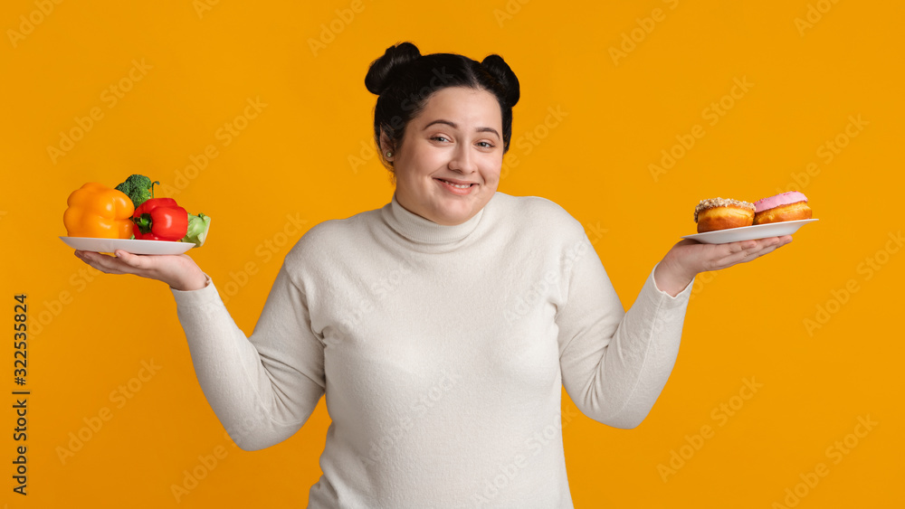 Positive plump woman holding plates with donuts and fresh organic vegetables Positive plump woman holding plates with donuts and fresh organic vegetables