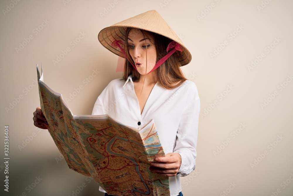 Young beautiful redhead woman wearing asian traditional hat holding city map scared in shock with a surprise face, afraid and excited with fear expression