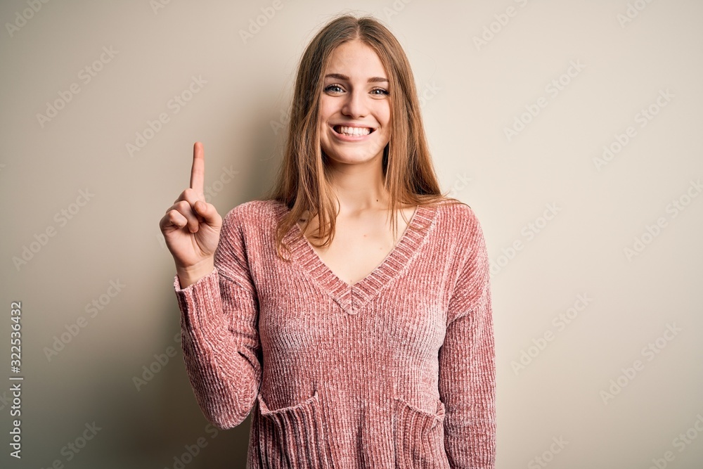 Young beautiful redhead woman wearing pink casual sweater over isolated white background showing and pointing up with finger number one while smiling confident and happy. Young beautiful redhead woman wearing pink casual sweater over isolated white background showing and pointing up with finger number one while smiling confident and happy.
