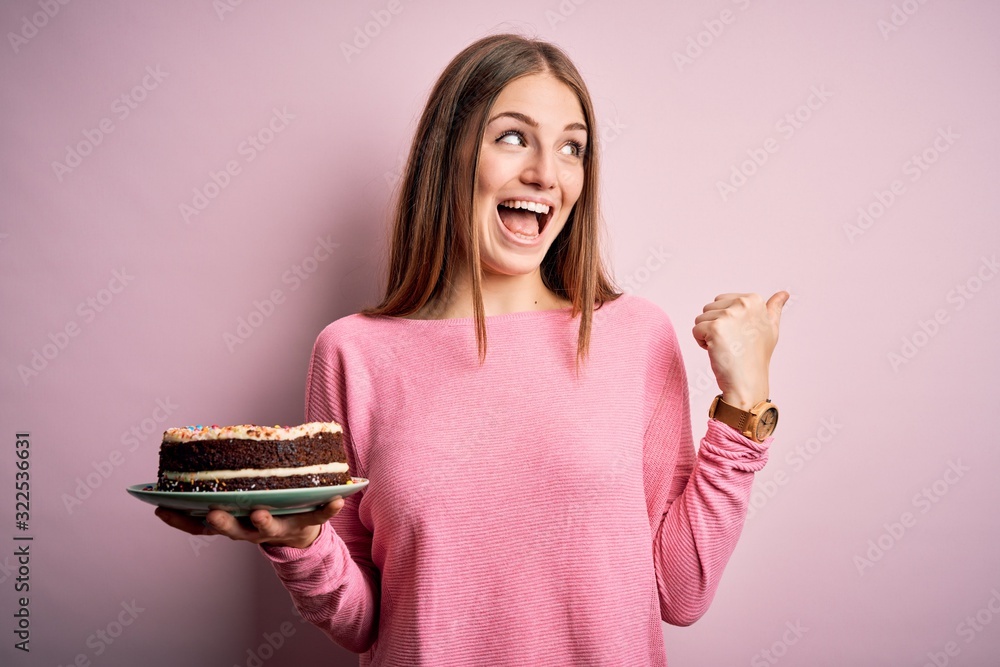 Young beautiful redhead woman holding birthday cake over isolated pink background pointing and showing with thumb up to the side with happy face smiling