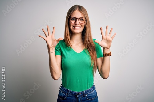 Young beautiful redhead woman wearing casual green t-shirt and glasses over white background showing and pointing up with fingers number ten while smiling confident and happy.