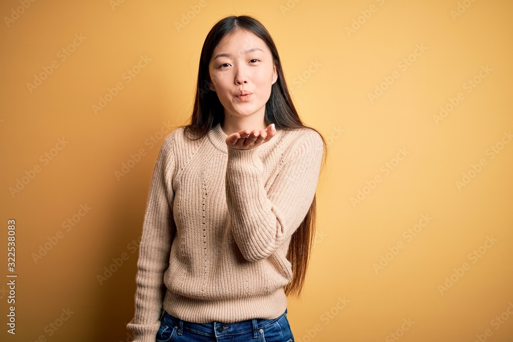 Young beautiful asian woman wearing casual sweater over yellow isolated background looking at the camera blowing a kiss with hand on air being lovely and sexy. Love expression. Young beautiful asian woman wearing casual sweater over yellow isolated background looking at the camera blowing a kiss with hand on air being lovely and sexy. Love expression.