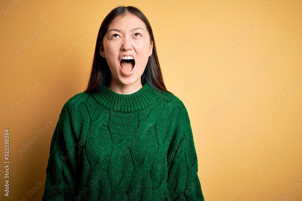 Fototapeta premium Young beautiful asian woman wearing green winter sweater over yellow isolated background angry and mad screaming frustrated and furious, shouting with anger. Rage and aggressive concept.