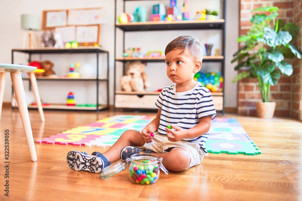 Beautiful toddler boy sitting on puzzle eating small colored chocolate balls at kindergarten