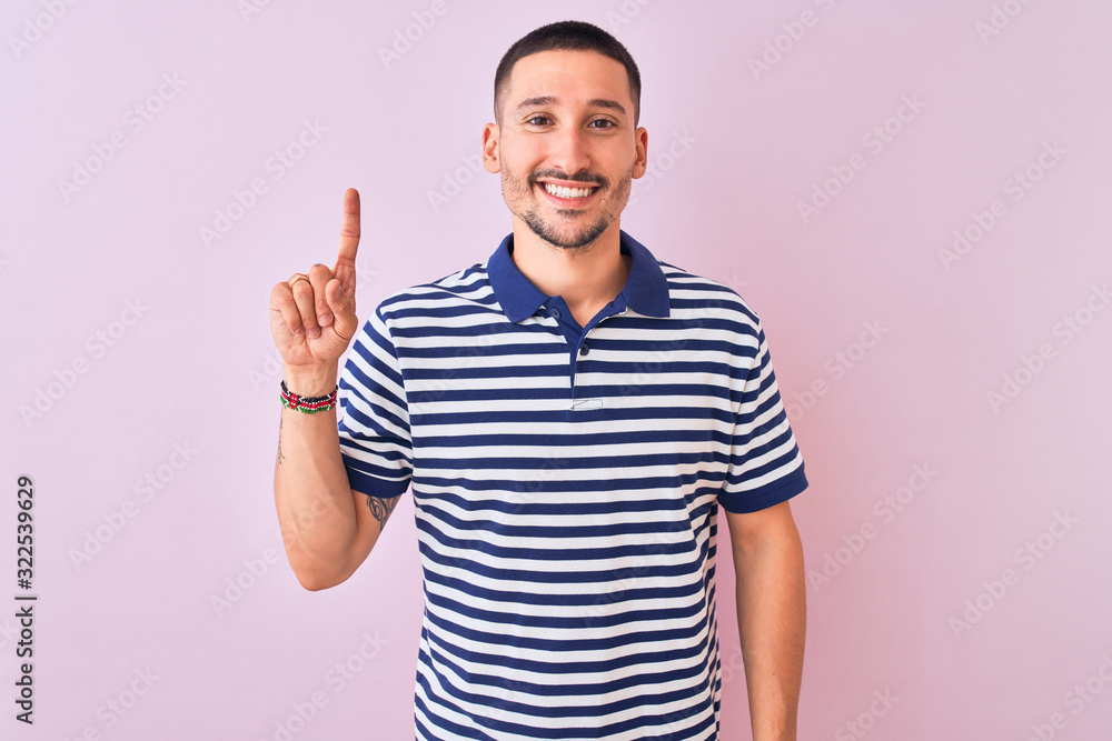 Young handsome man wearing nautical striped t-shirt over pink isolated background showing and pointing up with finger number one while smiling confident and happy. Young handsome man wearing nautical striped t-shirt over pink isolated background showing and pointing up with finger number one while smiling confident and happy.