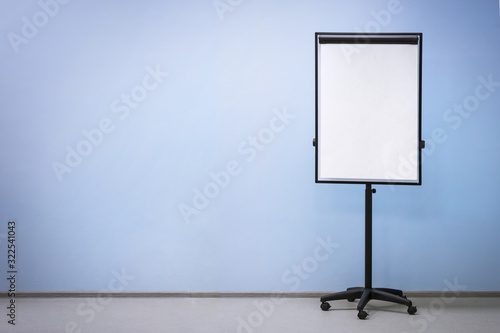 Flip chart board on wheels with a blank sheet of paper on a light blue wall background, copy space. Empty room for educational activities.