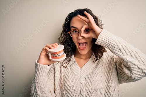 Young beautiful woman with curly hair holding plastic teeth with dental baces with happy face smiling doing ok sign with hand on eye looking through fingers photo