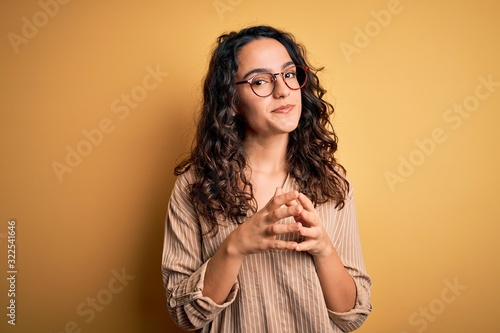 Beautiful woman with curly hair wearing striped shirt and glasses over yellow background Hands together and fingers crossed smiling relaxed and cheerful. Success and optimistic