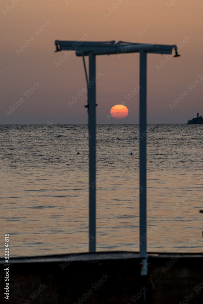 Focus on the round setting Sun. Blurred showers silhouettes at the beach at sunset in front of golden Ionian Sea water near beach. Dusk as seen from Ksamil, Albania, spring scenery evening.