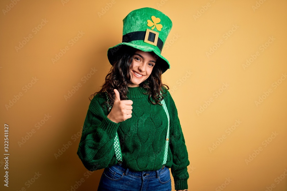 Beautiful curly hair woman wearing green hat with clover celebrating saint patricks day doing happy thumbs up gesture with hand. Approving expression looking at the camera showing success. Beautiful curly hair woman wearing green hat with clover celebrating saint patricks day doing happy thumbs up gesture with hand. Approving expression looking at the camera showing success.
