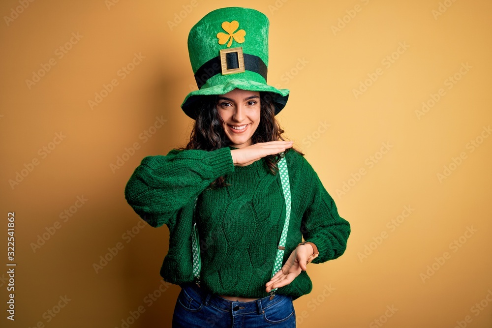Beautiful curly hair woman wearing green hat with clover celebrating saint patricks day gesturing with hands showing big and large size sign, measure symbol. Smiling looking at the camera. Measuring Beautiful curly hair woman wearing green hat with clover celebrating saint patricks day gesturing with hands showing big and large size sign, measure symbol. Smiling looking at the camera. Measuring
