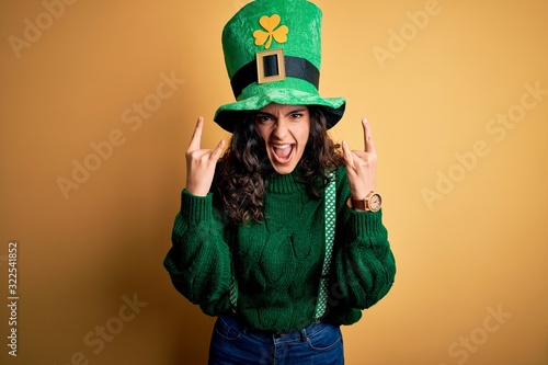 Canvas Print Beautiful curly hair woman wearing green hat with clover celebrating saint patricks day shouting with crazy expression doing rock symbol with hands up