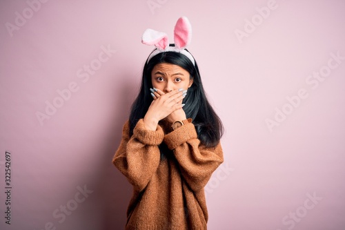 Young beautiful chinese woman wearing bunny ears standing over isolated pink background shocked covering mouth with hands for mistake. Secret concept.