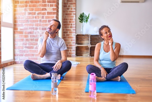 Middle age sporty couple sitting on mat doing stretching yoga exercise at gym with hand on chin thinking about question, pensive expression. Smiling with thoughtful face. Doubt concept.