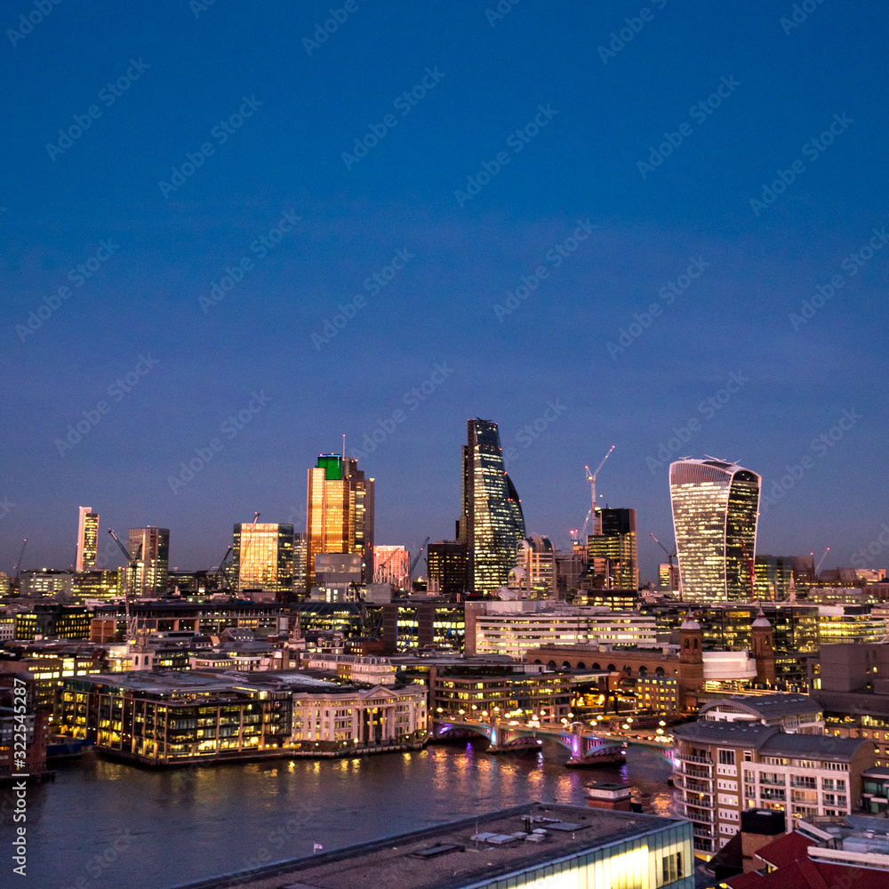 City of London at dusk. An elevated view of the illuminated financial district skyline fronted by the River Thames riverside.