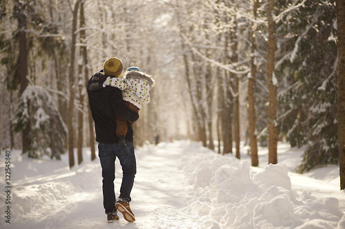 Young man with child on hands walking down the path in sunny winter park, snowy trees and snowbanks. View from behind