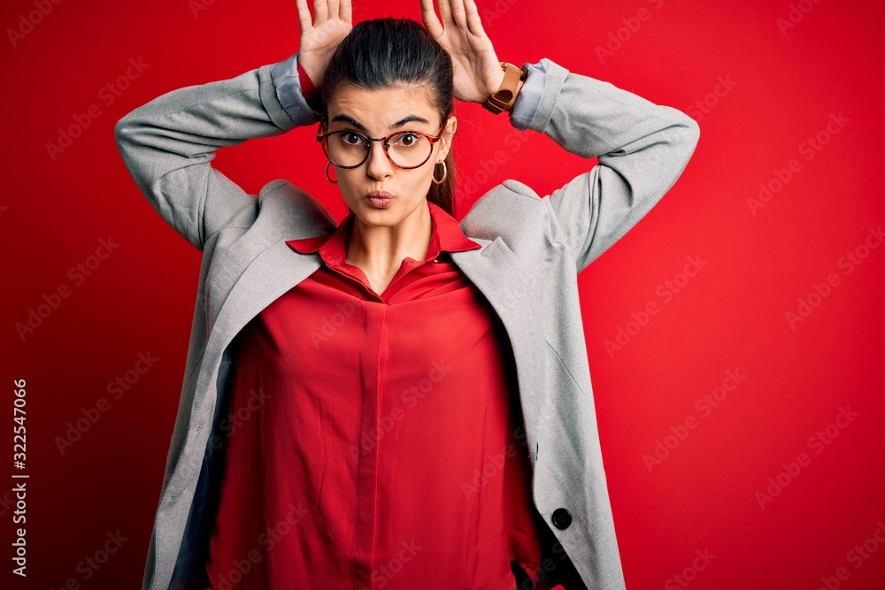 Young beautiful brunette businesswoman wearing jacket and glasses over red background Doing bunny ears gesture with hands palms looking cynical and skeptical. Easter rabbit concept. Young beautiful brunette businesswoman wearing jacket and glasses over red background Doing bunny ears gesture with hands palms looking cynical and skeptical. Easter rabbit concept.