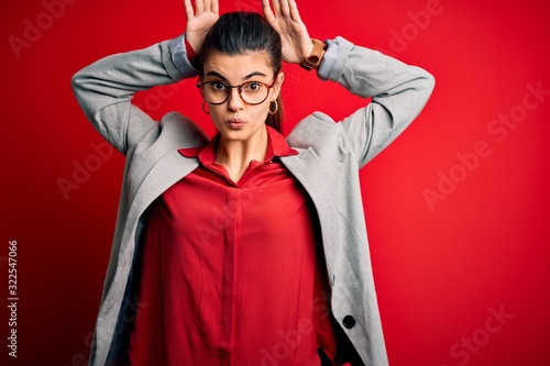 Young beautiful brunette businesswoman wearing jacket and glasses over red background Doing bunny ears gesture with hands palms looking cynical and skeptical. Easter rabbit concept.