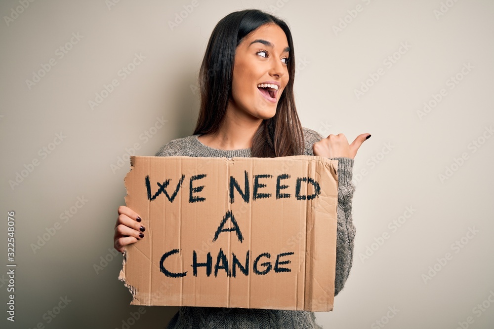 Young beautiful brunette activist woman protesting for a change holding banner pointing and showing with thumb up to the side with happy face smiling Young beautiful brunette activist woman protesting for a change holding banner pointing and showing with thumb up to the side with happy face smiling