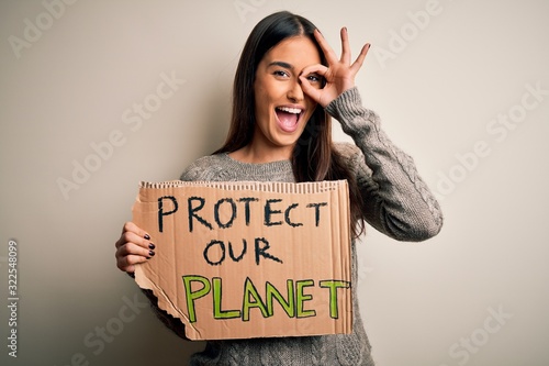 Young beautiful brunette activist woman protesting for protect our planet holding poster with happy face smiling doing ok sign with hand on eye looking through fingers © Krakenimages.com