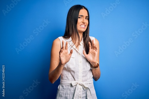 Young beautiful brunette woman wearing casual dress over isolated blue background disgusted expression, displeased and fearful doing disgust face because aversion reaction. With hands raised