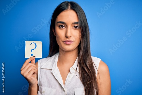 Young beautiful brunette woman holding paper with question mark symbol message with a confident expression on smart face thinking serious