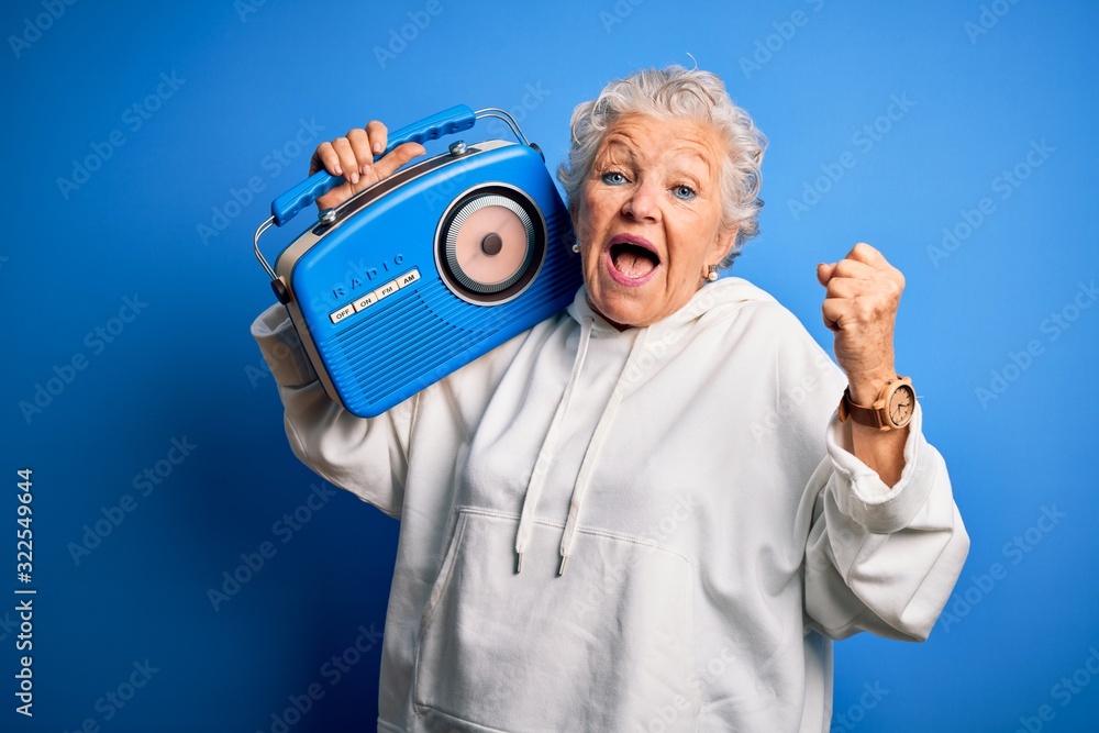 Naklejka premium Senior beautiful woman holding vintage radio standing over isolated blue background screaming proud and celebrating victory and success very excited, cheering emotion