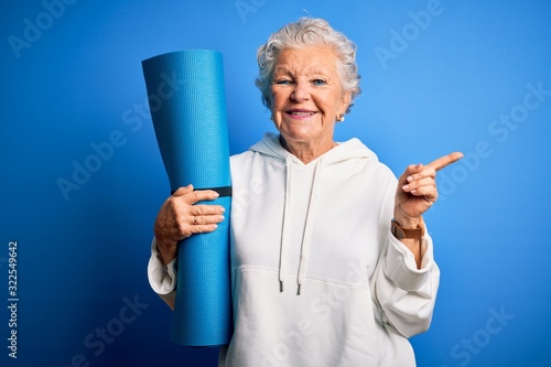 Fototapeta Naklejka Na Ścianę i Meble -  Senior beautiful sporty woman holding mat for yoga standing over isolated blue background very happy pointing with hand and finger to the side