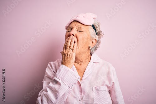 Senior beautiful woman wearing sleep mask and pajama over isolated pink background bored yawning tired covering mouth with hand. Restless and sleepiness. photo