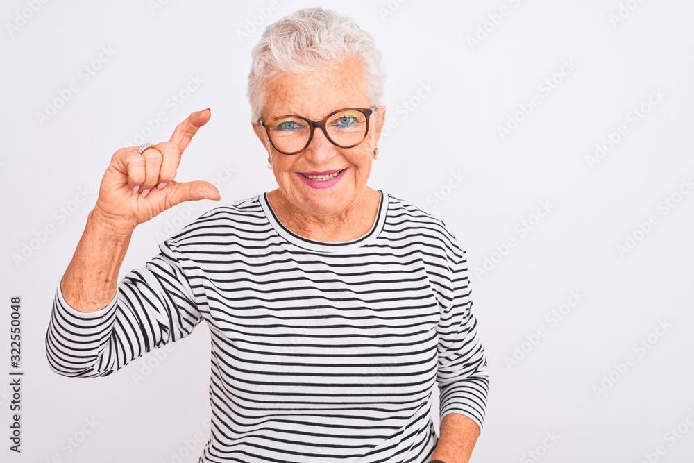Senior grey-haired woman wearing striped navy t-shirt glasses over isolated white background smiling and confident gesturing with hand doing small size sign with fingers looking and the camera. Senior grey-haired woman wearing striped navy t-shirt glasses over isolated white background smiling and confident gesturing with hand doing small size sign with fingers looking and the camera.