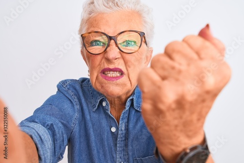 Grey-haired woman wearing denim shirt and glasses make selfie over isolated white background annoyed and frustrated shouting with anger, crazy and yelling with raised hand, anger concept