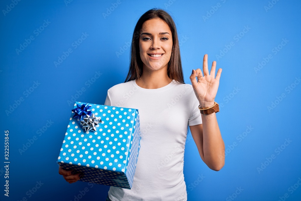 Young beautiful brunette woman holding birthday gift over isolated blue background doing ok sign with fingers, excellent symbol Young beautiful brunette woman holding birthday gift over isolated blue background doing ok sign with fingers, excellent symbol