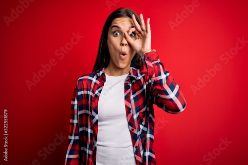 Young beautiful brunette woman wearing casual shirt standing over isolated red background doing ok gesture shocked with surprised face, eye looking through fingers. Unbelieving expression. © Krakenimages.com