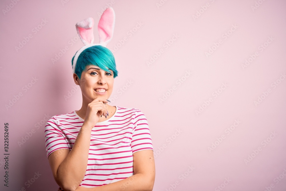 Young woman with fashion blue hair wearing easter rabbit ears over pink background looking confident at the camera with smile with crossed arms and hand raised on chin. Thinking positive.