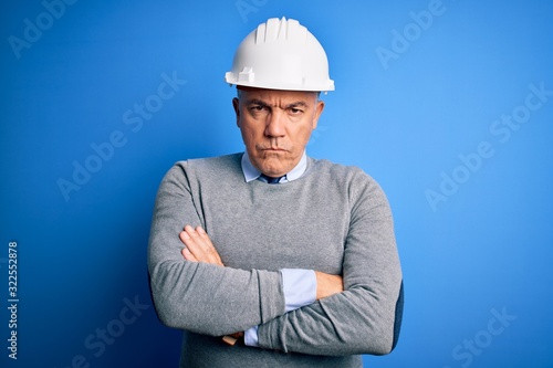 Middle age handsome grey-haired engineer man wearing safety helmet over blue background skeptic and nervous, disapproving expression on face with crossed arms. Negative person.