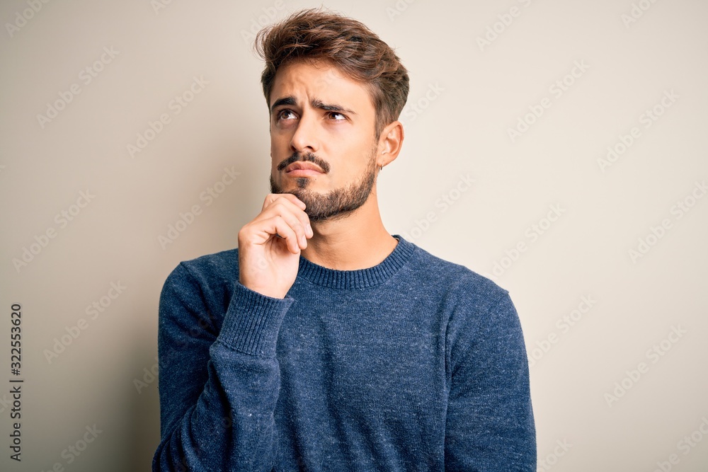 Young handsome man with beard wearing casual sweater standing over white background with hand on chin thinking about question, pensive expression. Smiling with thoughtful face. Doubt concept. Young handsome man with beard wearing casual sweater standing over white background with hand on chin thinking about question, pensive expression. Smiling with thoughtful face. Doubt concept.