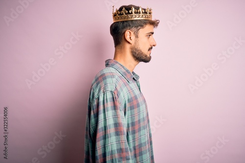 Young man with beard wearing golden crown of king standing over isolated pink background looking to side, relax profile pose with natural face with confident smile.
