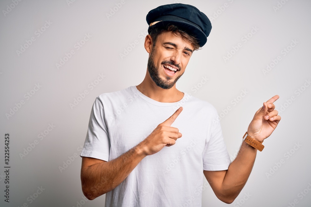 Young driver man with beard wearing hat standing over isolated white background smiling and looking at the camera pointing with two hands and fingers to the side. Young driver man with beard wearing hat standing over isolated white background smiling and looking at the camera pointing with two hands and fingers to the side.