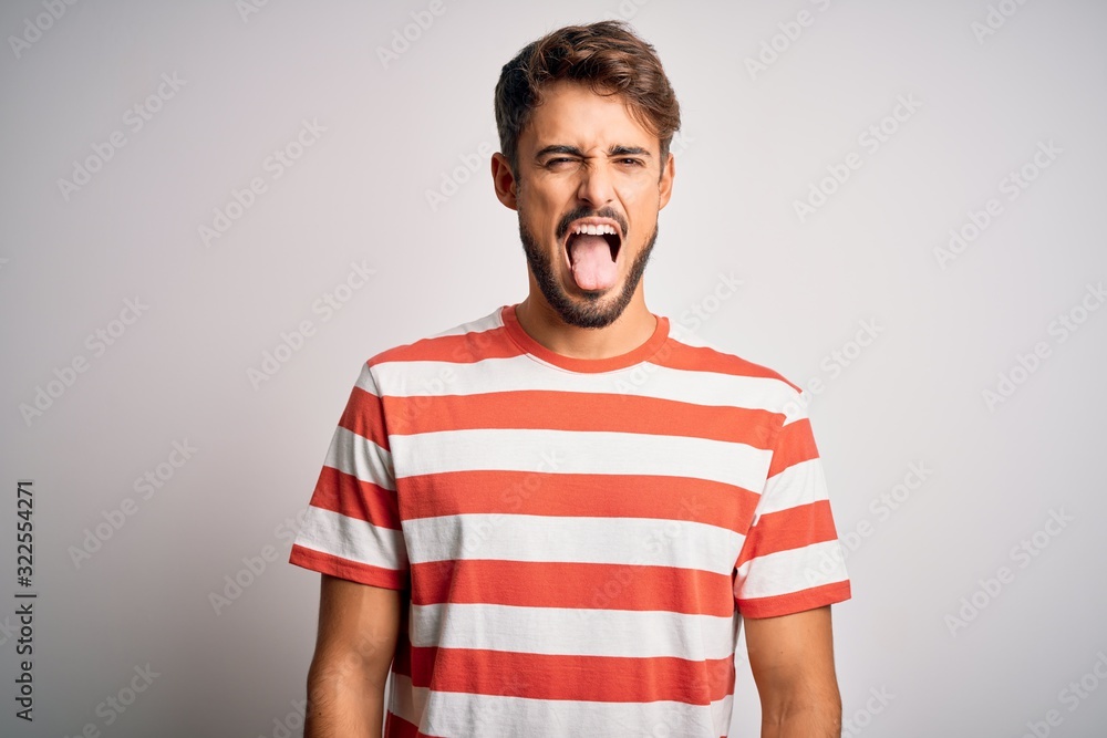 Young handsome man with beard wearing striped t-shirt standing over white background sticking tongue out happy with funny expression. Emotion concept. Young handsome man with beard wearing striped t-shirt standing over white background sticking tongue out happy with funny expression. Emotion concept.