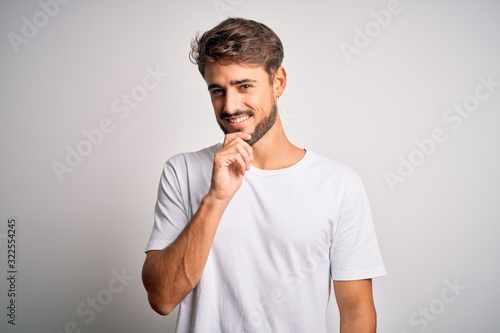 Young handsome man with beard wearing casual t-shirt standing over white background looking confident at the camera with smile with crossed arms and hand raised on chin. Thinking positive.