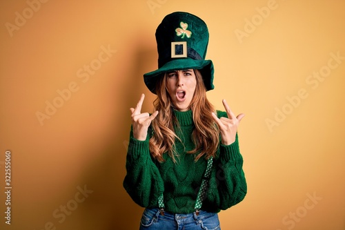 Canvas Print Beautiful brunette woman wearing green hat with clover celebrating saint patricks day shouting with crazy expression doing rock symbol with hands up